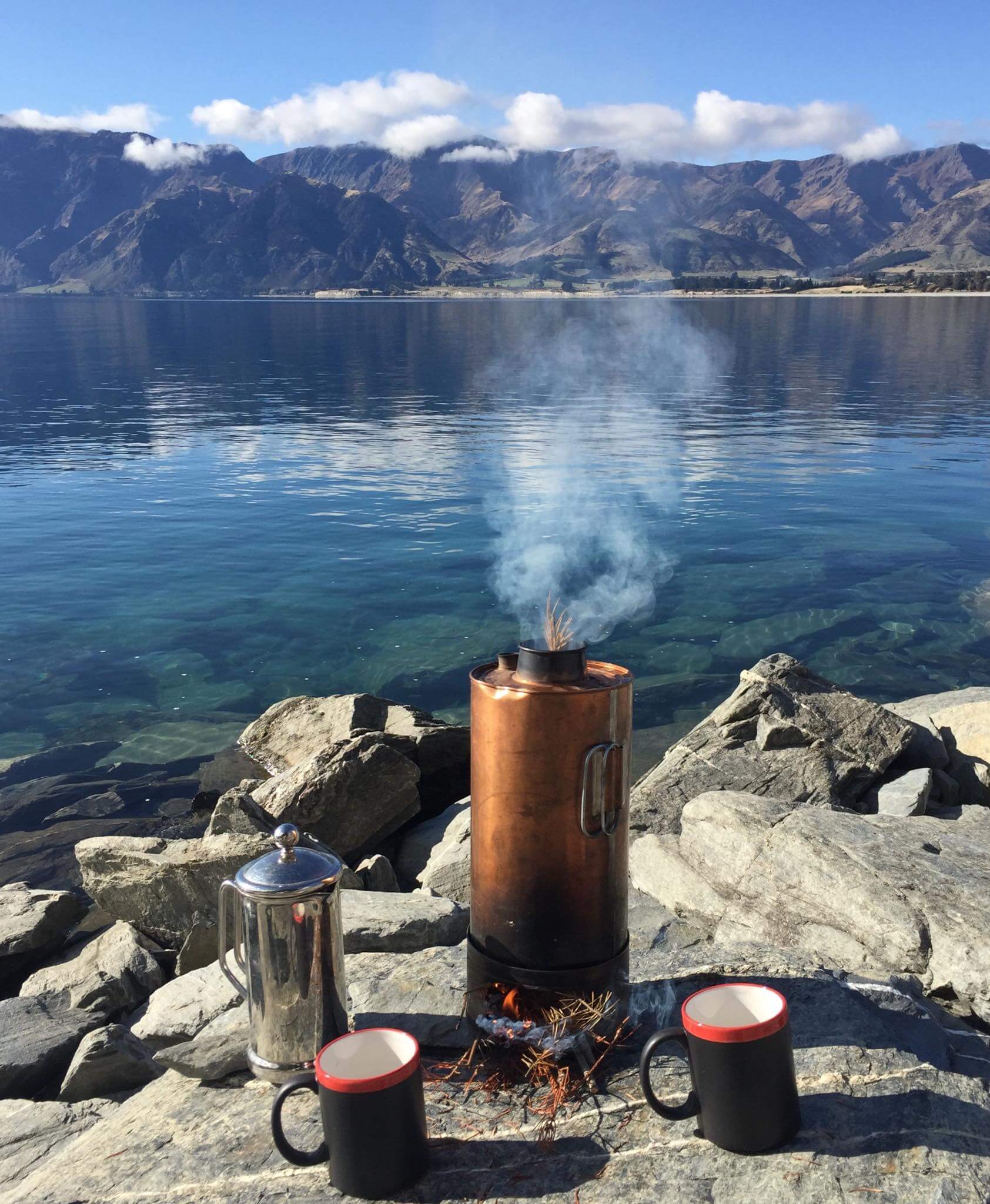 Why the Thermette Kettle is the Ultimate Outdoor Icon on a lakeside with coffee mugs and majestic mountains in the background.