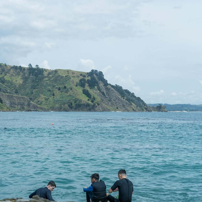 Three people in wetsuits fishing near water with rocky shore and hills in the background.