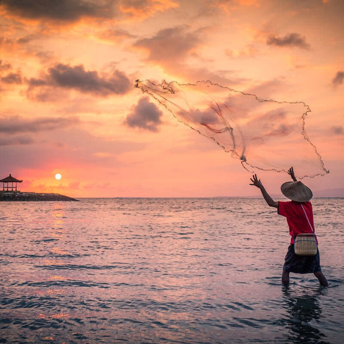 Person casting a net in the ocean at sunset with a pavilion in the background.