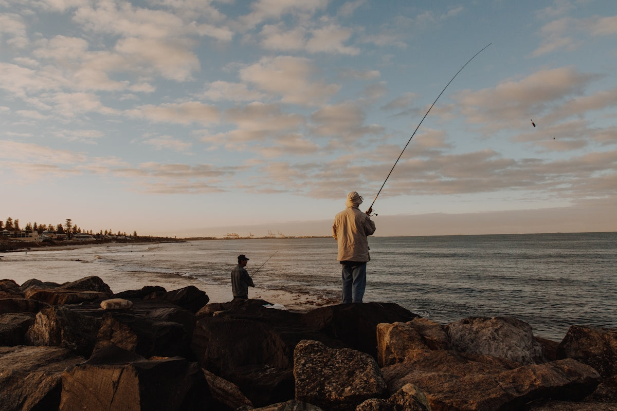 man in white shirt and black pants holding fishing rod standing on rock near sea during