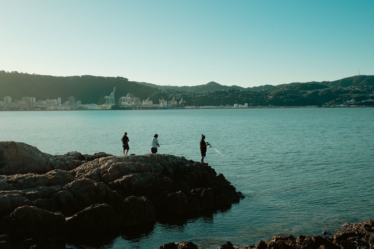 Three people fishing on rocks by a calm bay.
